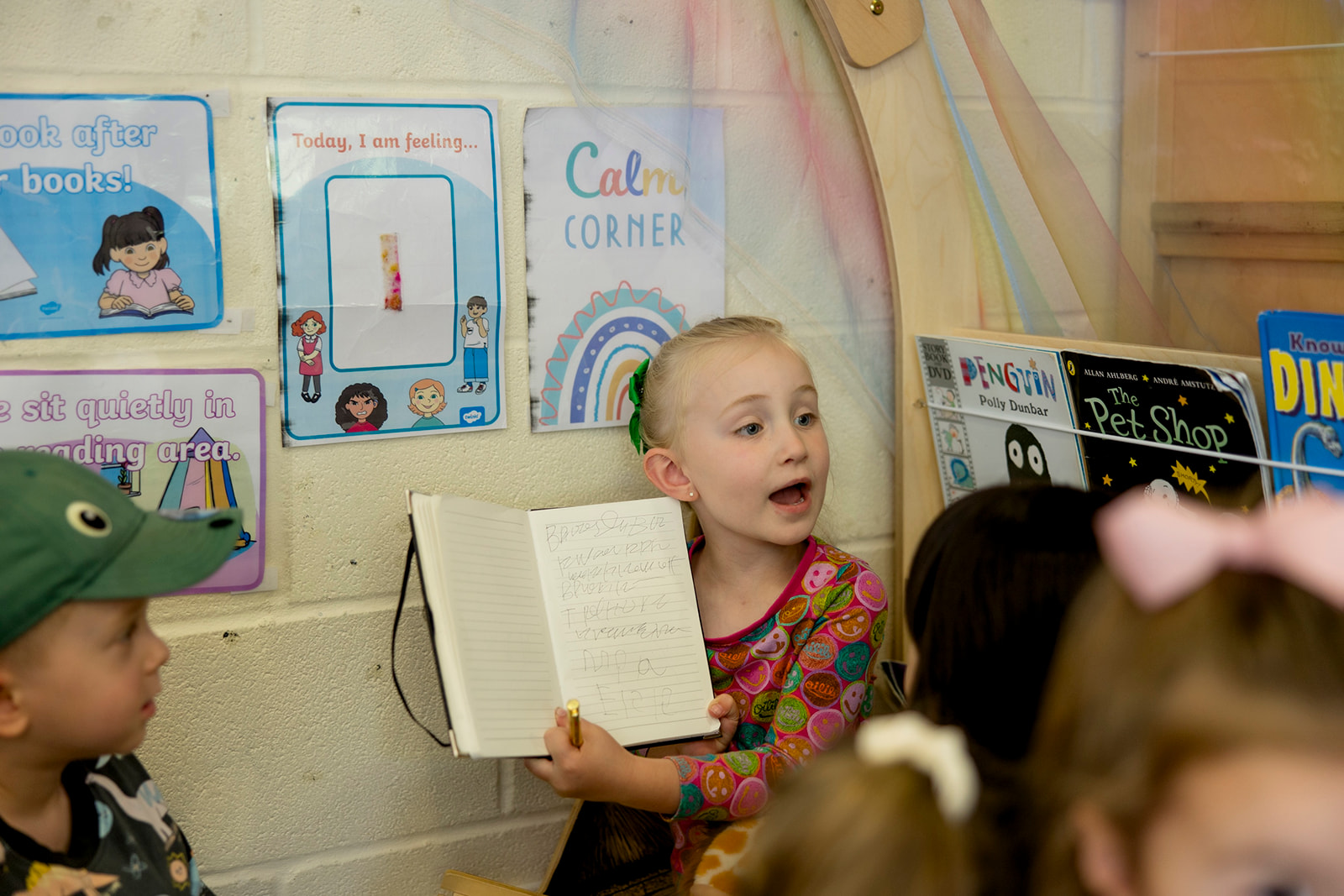 A girl in the book corner reading aloud from her notebook to other children