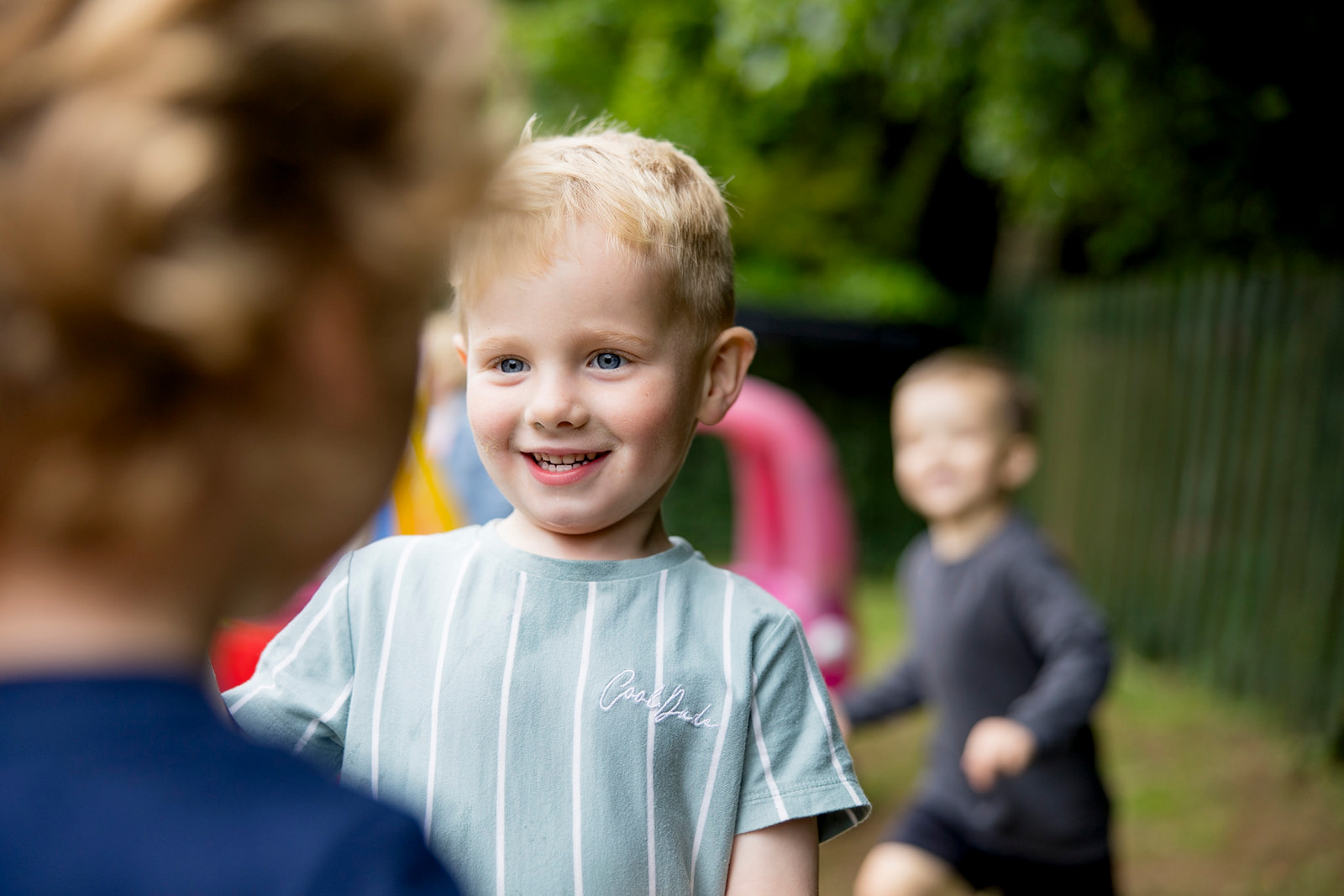 A smiling boy in the preschool garden with children playing behind him