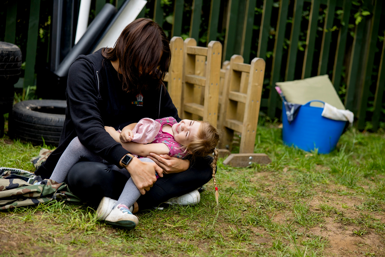 A member of staff sitting on the grass cuddling a laughing child in the preschool garden